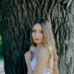 A young woman in a white dress stands elegantly by a large tree outdoors, capturing a serene moment.