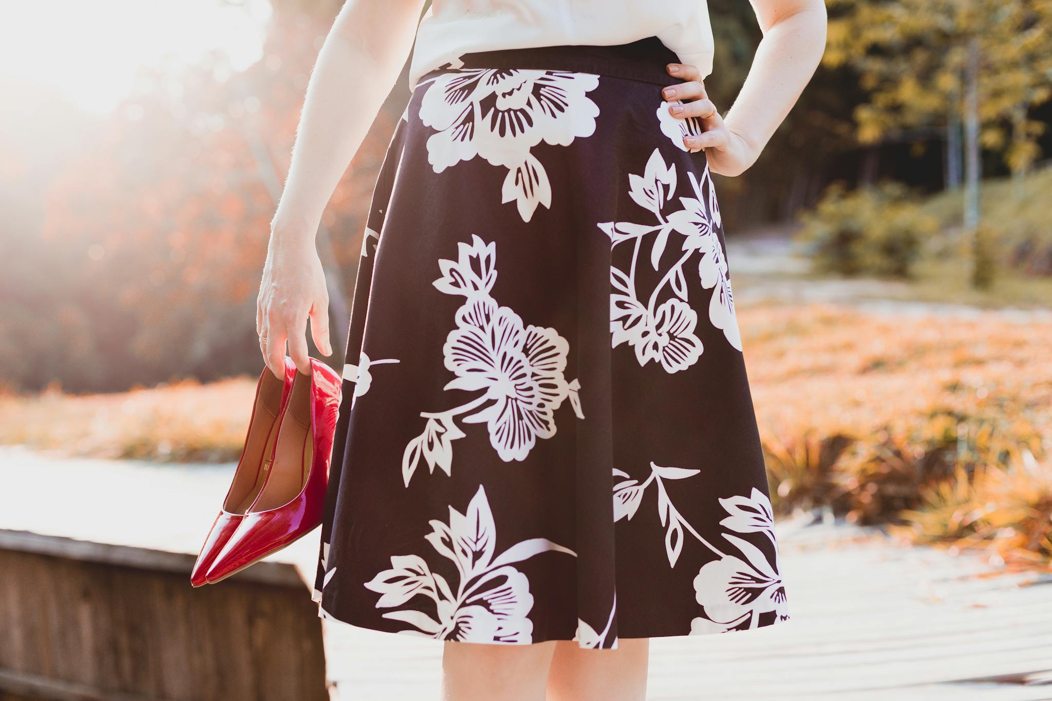 Woman holding red heels, wearing a floral skirt, enjoying a sunny day outdoors.